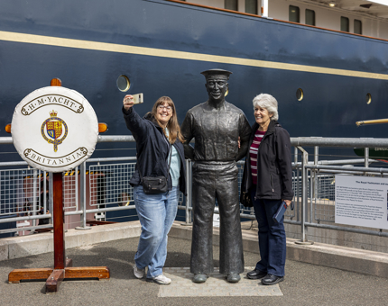 Two visitors standing next to a statue of of Norrie, an ex-Royal Yachtsman, taking a selfie.