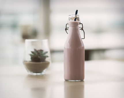 A strawberry milkshake in a small bottle with a straw. There is a small vase on the table with a plant in it.