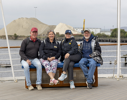 Four visitors sitting on a bench on the Verandah Deck. Behind them is water and the working port that has a mound of sand on it.