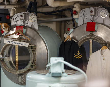 The Laundry Room on Britannia. There are two uniforms hanging from one machine. The other machine has a small corgi plush toy on it.
